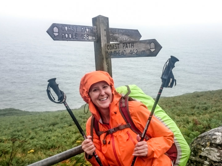 Gilly in an orange waterproof with walking poles beside a Coast Path sign